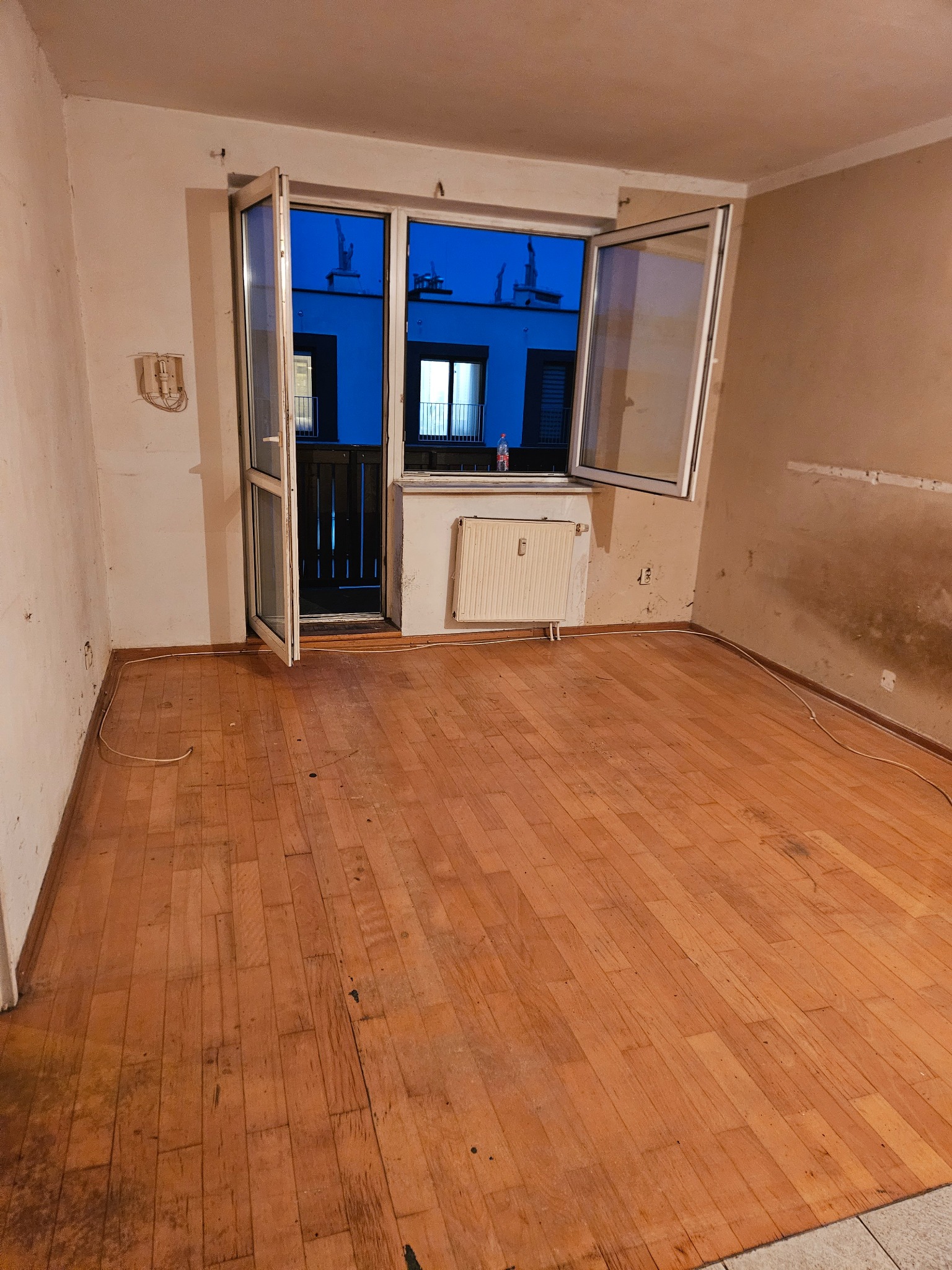 Puste, wysprzątane mieszkanie z drewnianą podłogą i otwartym oknem balkonowym. Widoczne ślady po remoncie na ścianach. Efekt po opróżnianiu.