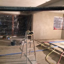 Mar-Bud Marcin Wąs - Wnętrze w trakcie tynkowania: ściany pokryte świeżym tynkiem, okna zabezpieczone folią, widoczne rury instalacyjne i drabina malarska. Prace wykończeniowe w toku.