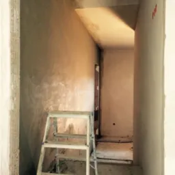 Wnętrze w trakcie prac wykończeniowych: ściany pokryte świeżo nałożonym tynkiem, drabina malarska w rogu, widoczne ślady po instalacjach elektrycznych.