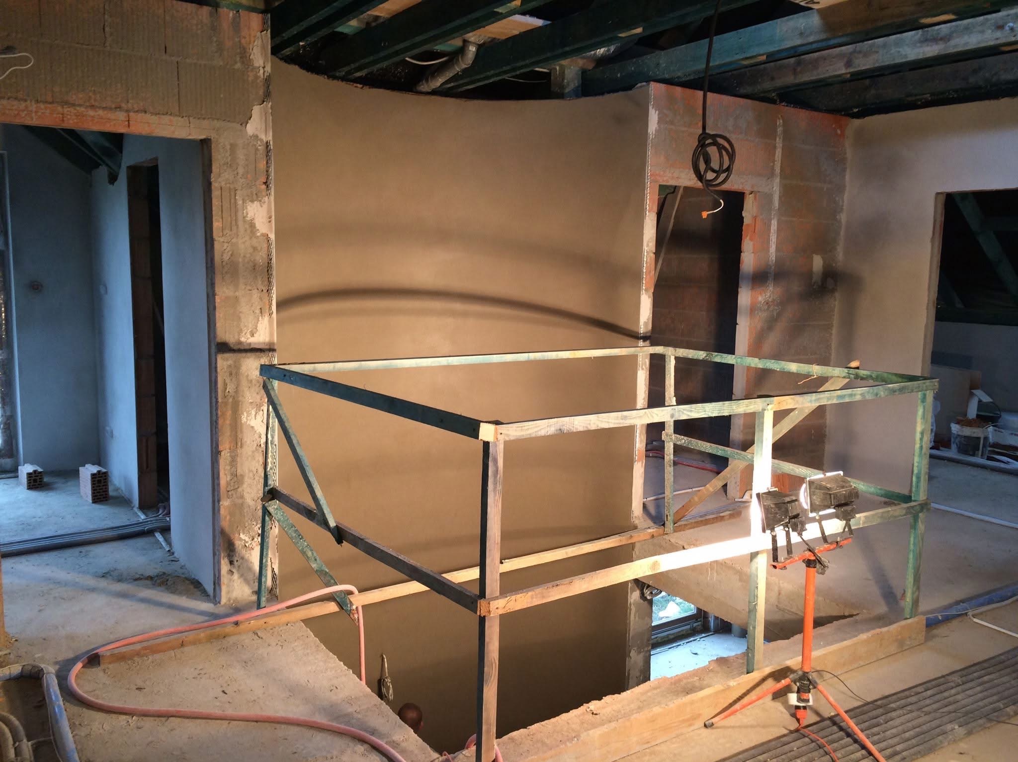 Wnętrze w trakcie prac wykończeniowych: ściany pokryte świeżym tynkiem, widoczne elementy konstrukcyjne, prowizoryjne barierki i lampa budowlana na trójnogu.