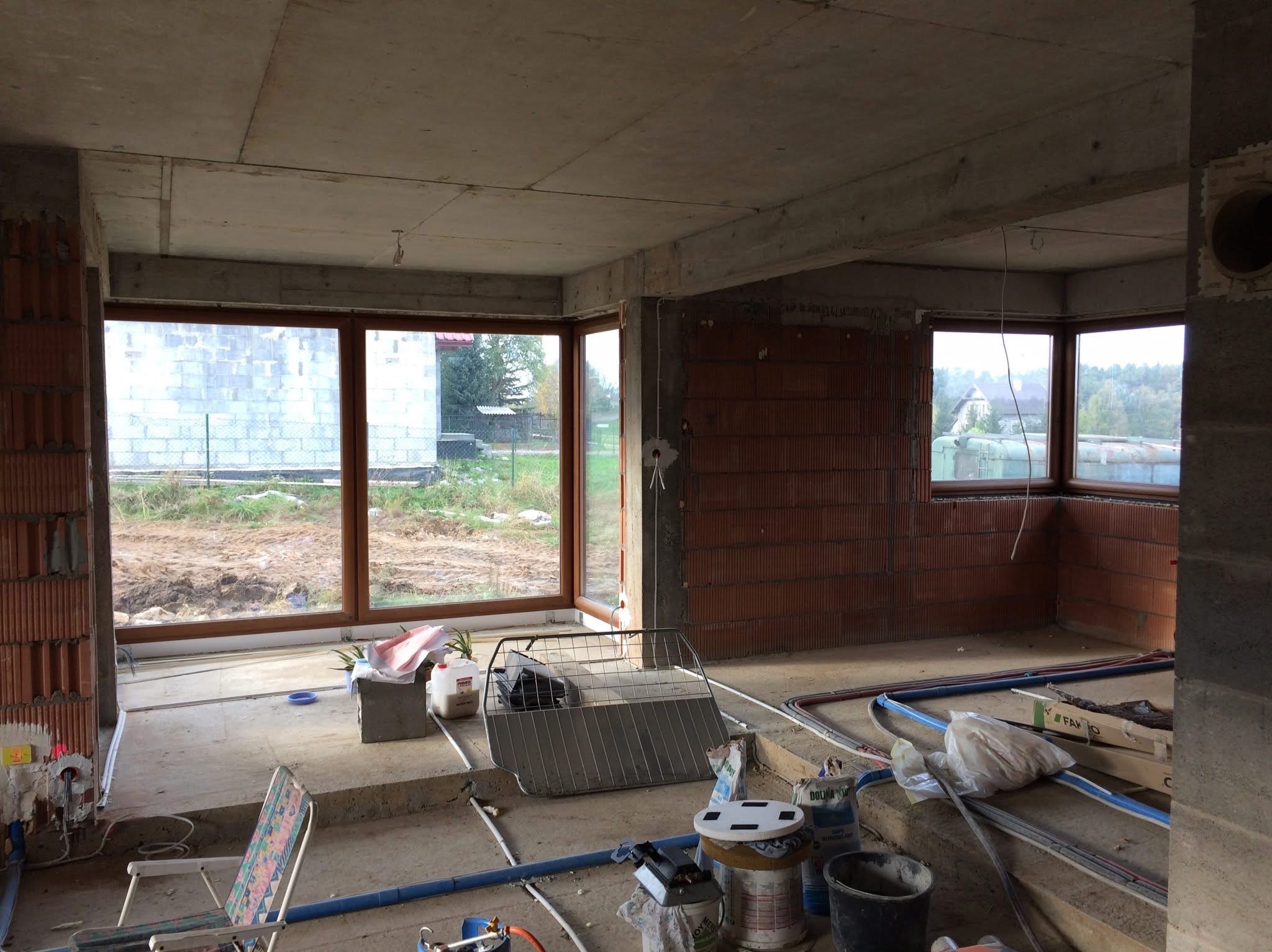 Surowy stan wnętrza domu w trakcie budowy, z widocznymi instalacjami podłogowymi, ceglanymi ścianami i dużymi oknami z widokiem na zewnątrz.