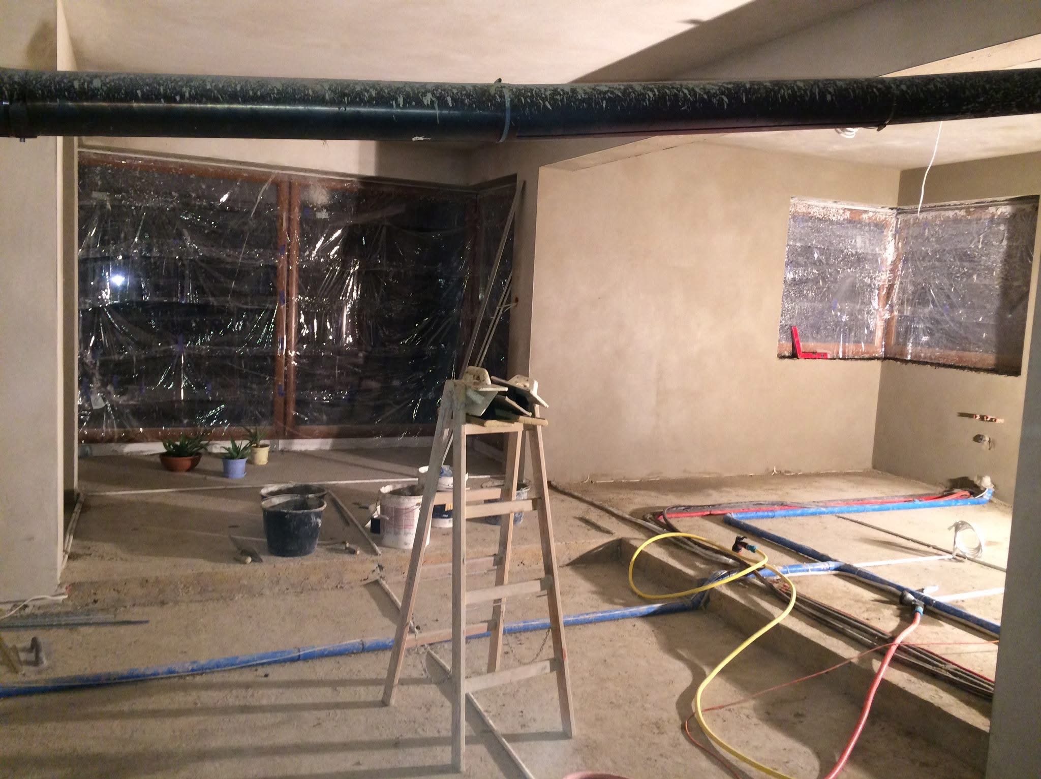 Wnętrze w trakcie tynkowania: ściany pokryte świeżym tynkiem, okna zabezpieczone folią, widoczne rury instalacyjne i drabina malarska. Prace wykończeniowe w toku.