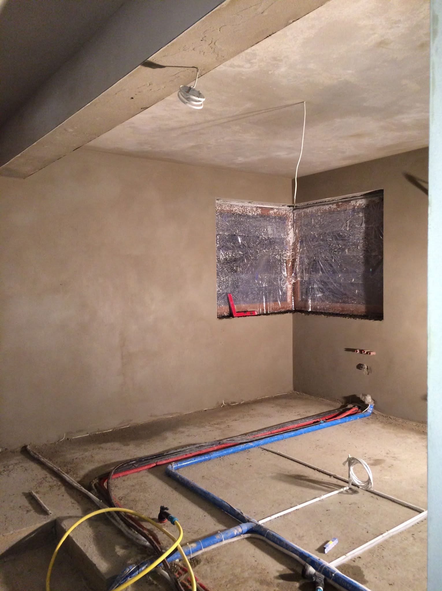 Wnętrze po tynkowaniu ścian, z zabezpieczonym oknem folią i widocznymi instalacjami na podłodze. Ściany mają surowy, jasnobrązowy odcień.