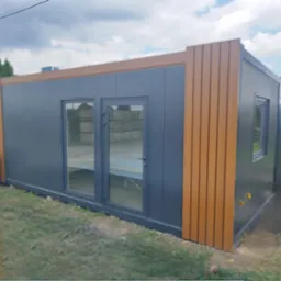 Nowoczesny pawilon z płyt warstwowych w kolorze grafitowym, z brązowymi akcentami wykończeniowymi i przeszklonymi drzwiami, na tle zieleni.