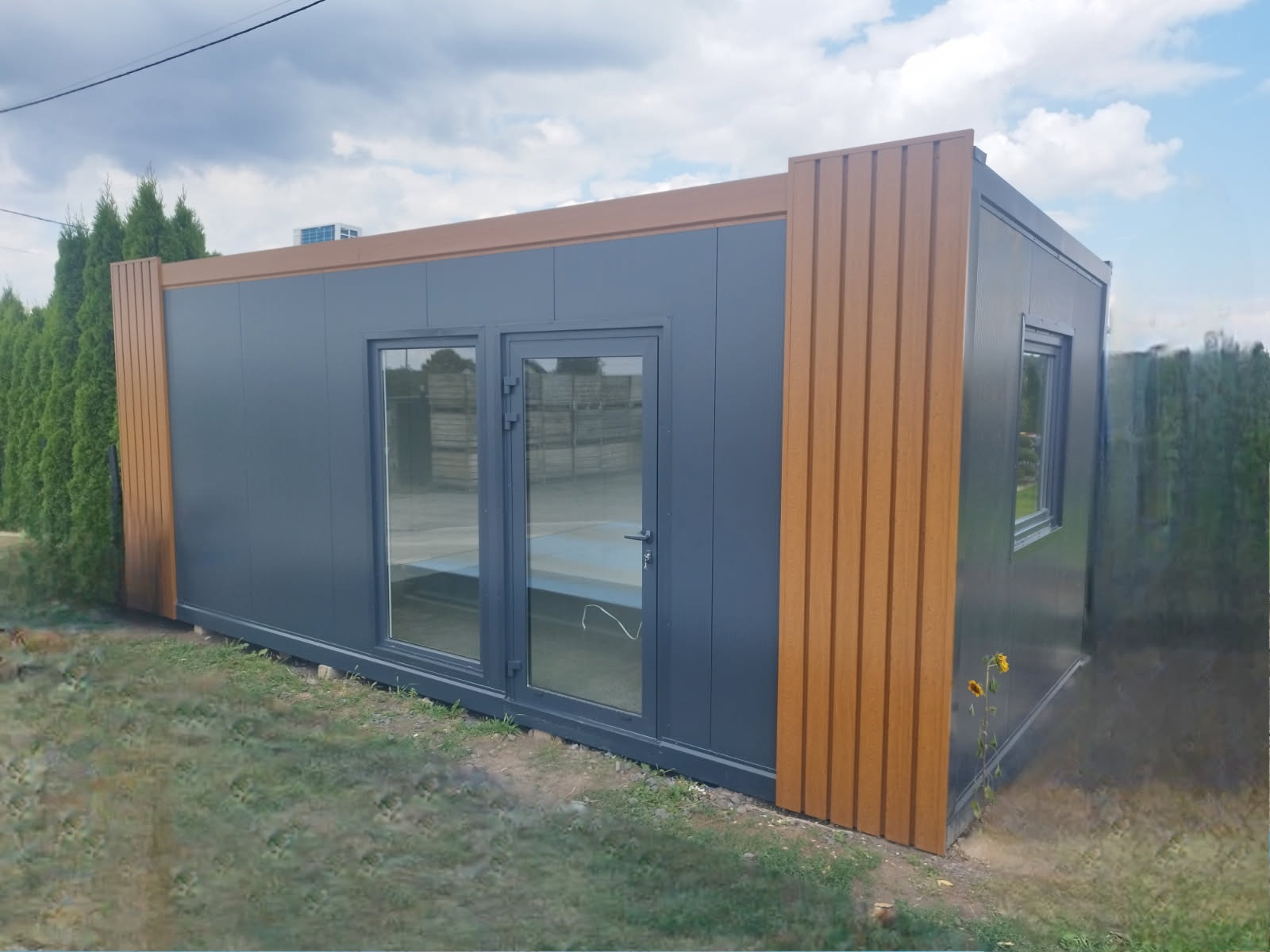 Nowoczesny pawilon z płyt warstwowych w kolorze grafitowym, z brązowymi akcentami wykończeniowymi i przeszklonymi drzwiami, na tle zieleni.