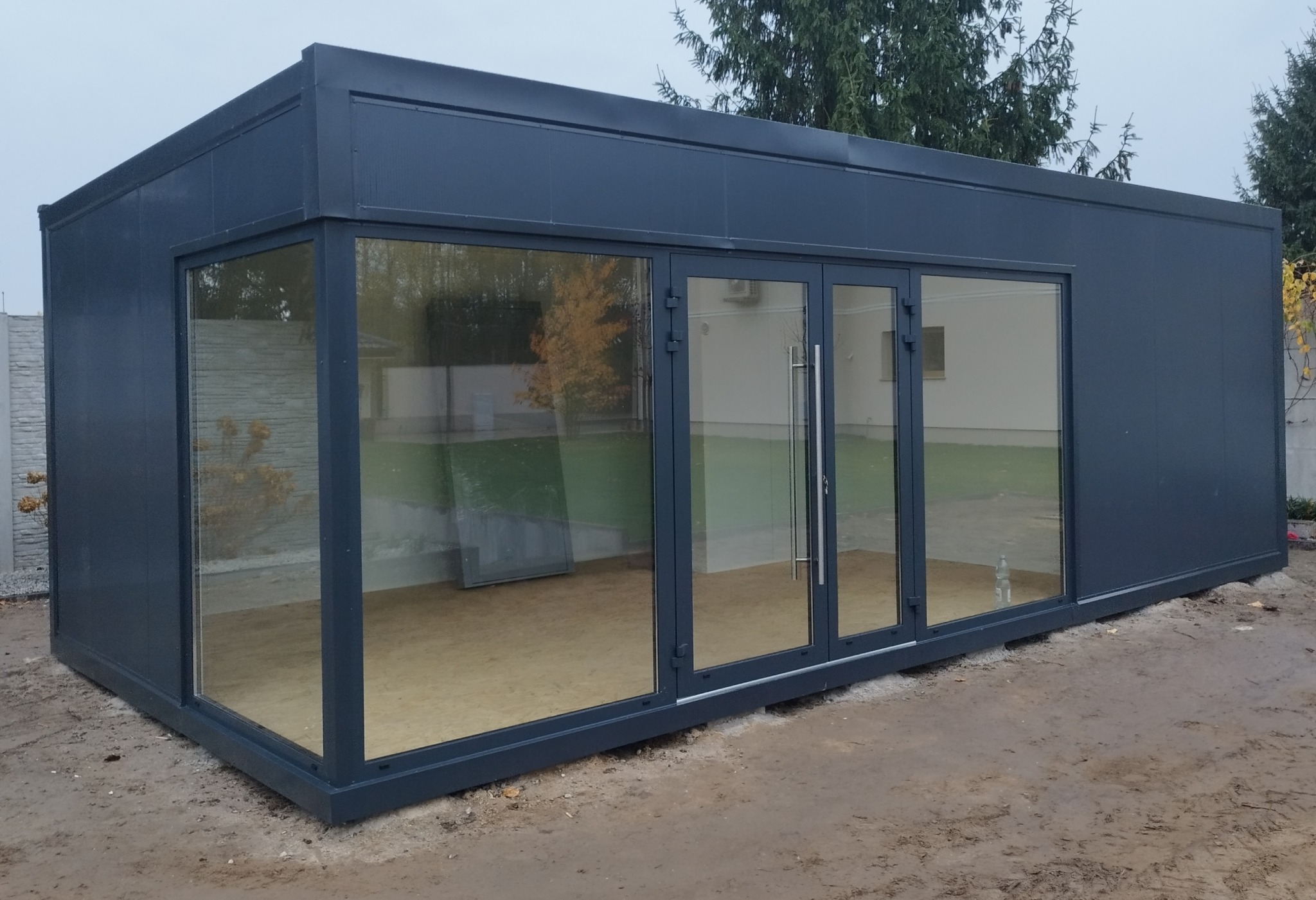 Szary pawilon z płyt warstwowych z dużymi oknami i drzwiami, na zewnątrz widoczne drzewa i trawa. Konstrukcja modułowa na ziemi, w tle fragment ogrodzenia.