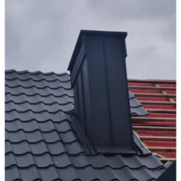 Czarny komin na dachu z blachy w odcieniach szarości, częściowo widoczna więźba dachowa w kolorze czerwonym. Ujęcie z dołu, pochmurne niebo w tle.