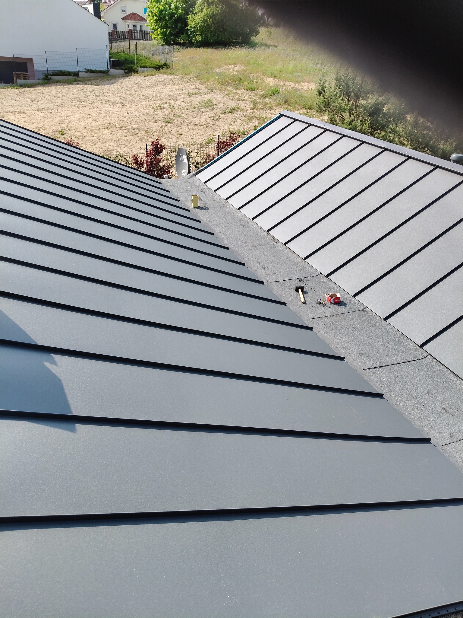 Szary dach z blachy na rąbek stojący, widok z góry, z narzędziami dekarskimi na kalenicy. W tle teren budowy i domy.