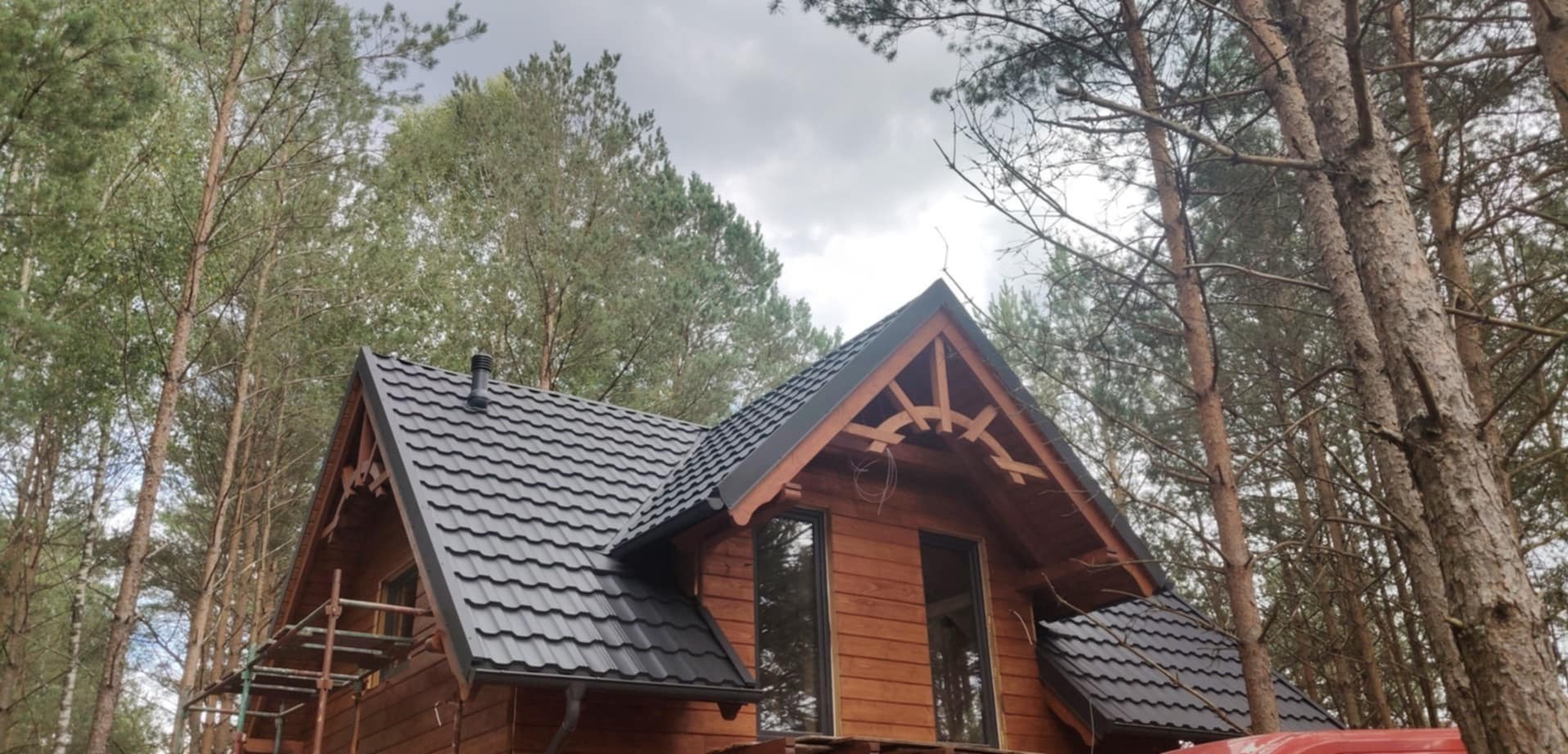 Drewniany dom z ciemną, metalową dachówką w lesie, z widocznymi rusztowaniami sugerującymi prace wykończeniowe. Dekoracyjne elementy drewniane na szczycie dachu.