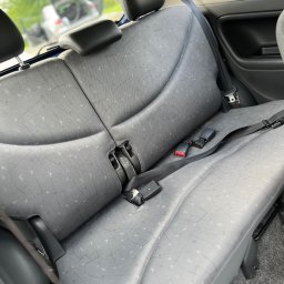 JTB LUX - Szare, tekstylne siedzenie samochodowe z zagłówkami i pasami bezpieczeństwa. W tle widoczny bagażnik i zieleń. Ujęcie z wnętrza pojazdu.