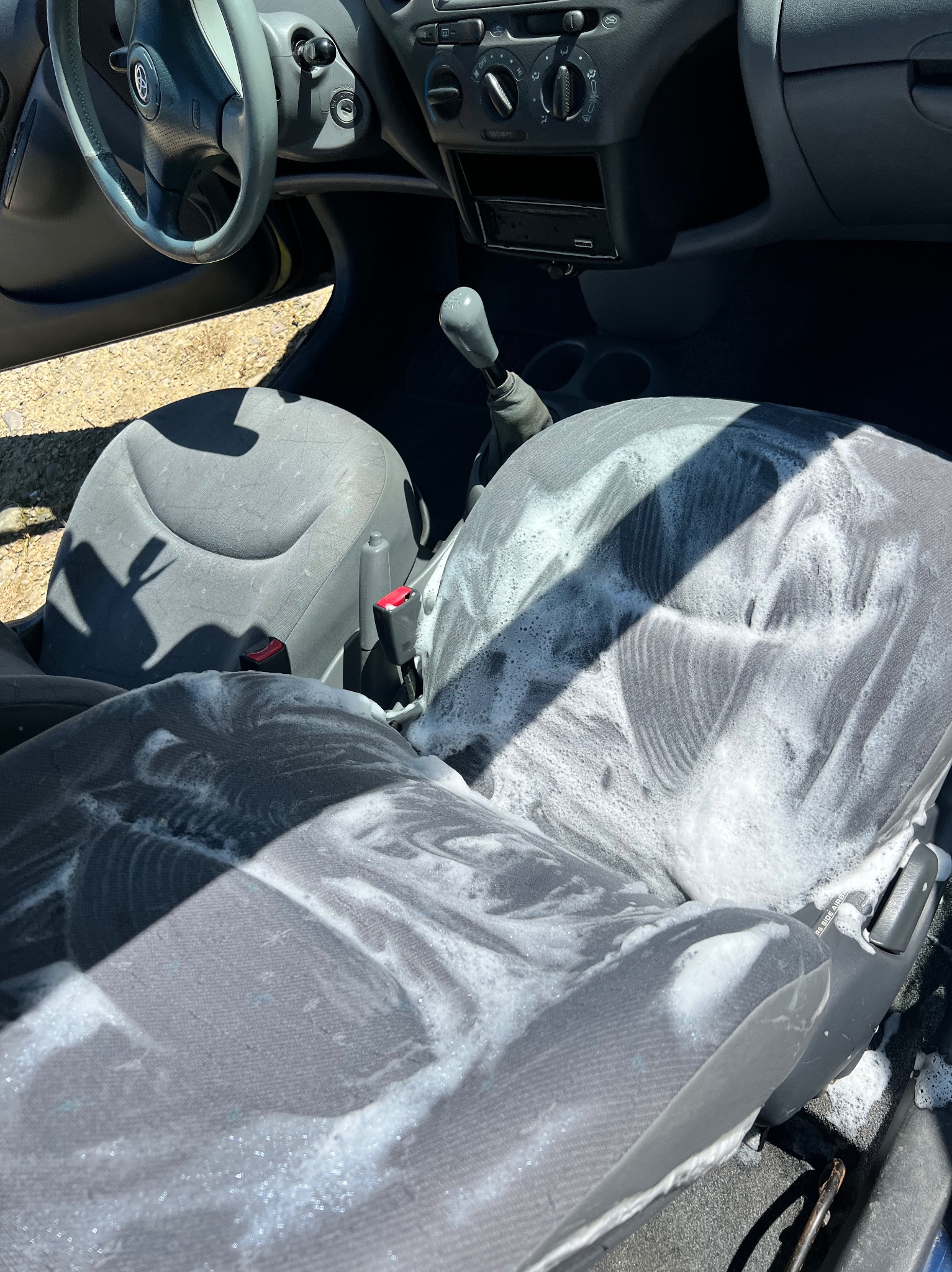 Piana na szarych fotelach samochodowych podczas czyszczenia. Widoczne wnętrze pojazdu i fragment kierownicy. Jasne oświetlenie.