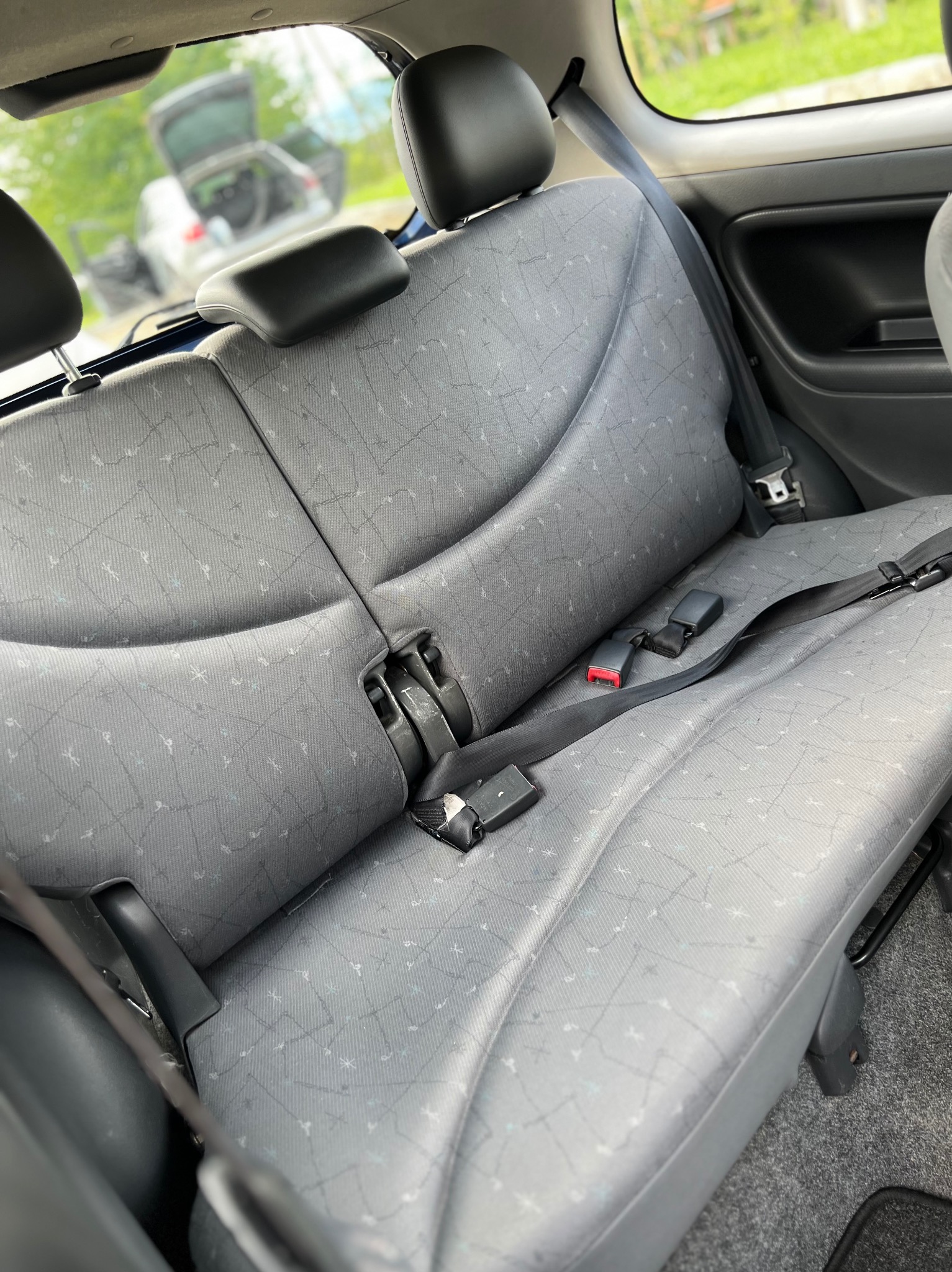 Szare, tekstylne siedzenie samochodowe z zagłówkami i pasami bezpieczeństwa. W tle widoczny bagażnik i zieleń. Ujęcie z wnętrza pojazdu.
