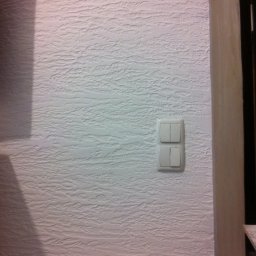 Ekipa malarska - Biała ściana z fakturą, na której zamontowany jest podwójny włącznik światła. Fragment futryny drzwi po prawej stronie. Delikatny cień w lewym górnym rogu.