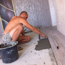 Ekipa malarska - Mężczyzna w szortach układa podłogowe płytki imitujące drewno w nowym pomieszczeniu. Widoczny poziomica, wiadro z zaprawą i krzyżyki dystansowe.