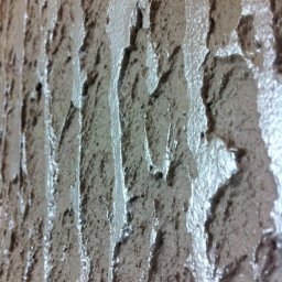 Ekipa malarska - Ściana pokryta świeżo nałożoną, grubą warstwą szarej gładzi strukturalnej z wyraźnymi, pionowymi śladami narzędzia, tworzącymi unikalną fakturę.