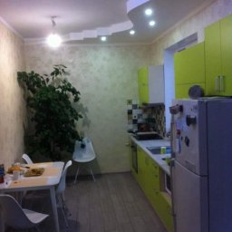 Ekipa malarska - Jasna kuchnia z zielonymi szafkami i dekoracyjnym sufitem. Widoczne panele podłogowe, stół z krzesłami i roślina doniczkowa w rogu pomieszczenia.