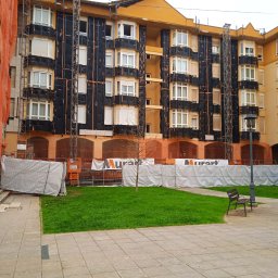 Ekipa malarska - Ocieplanie budynku wielorodzinnego z zamontowanym rusztowaniem i siatką zabezpieczającą. Widoczne płyty izolacyjne na elewacji w kolorze czarnym.