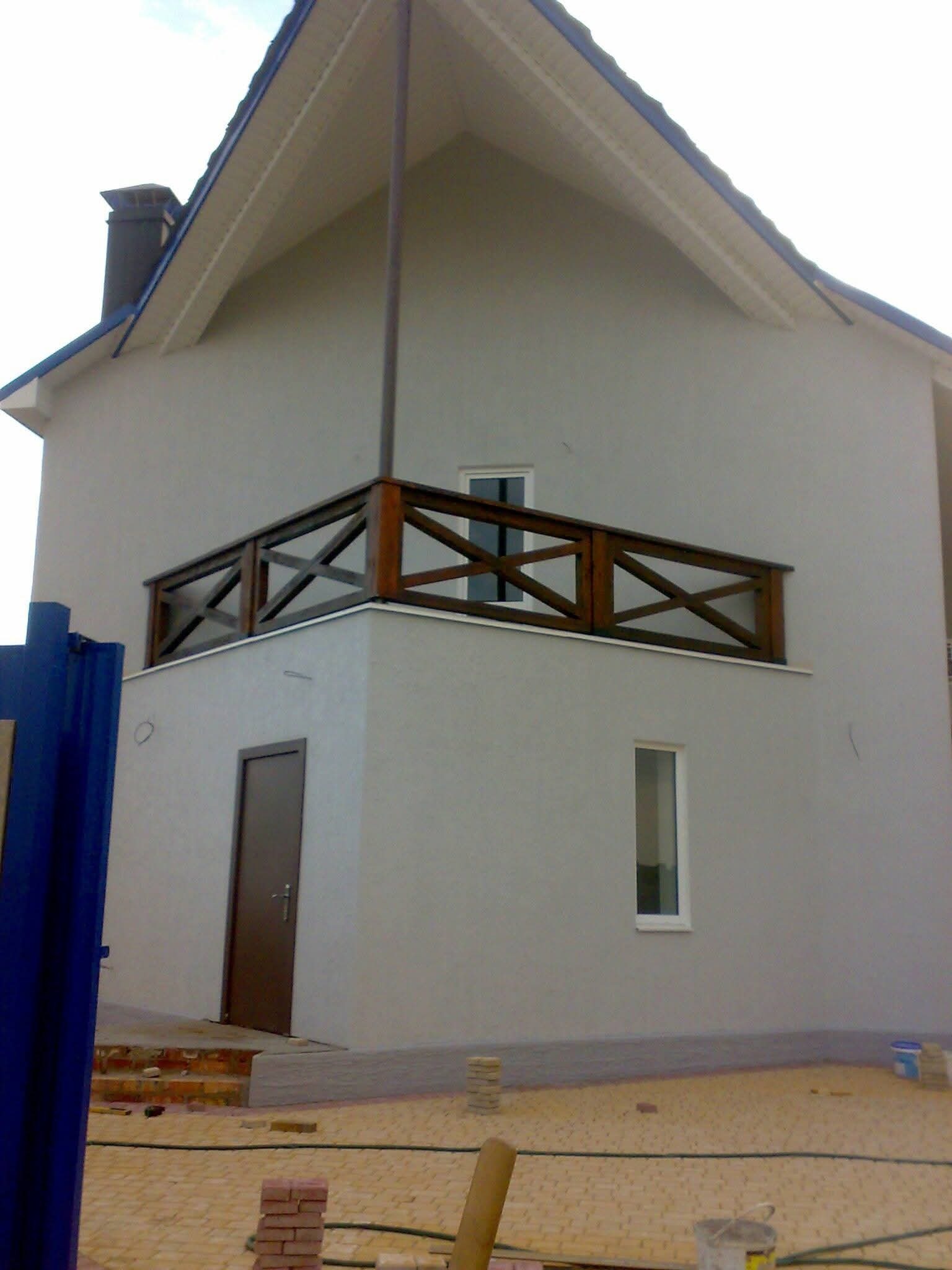 Szary dom z drewnianą balustradą balkonu, widok z zewnątrz. W tle błękitne niebo i fragment dachu. Na ziemi kostka brukowa i materiały budowlane.