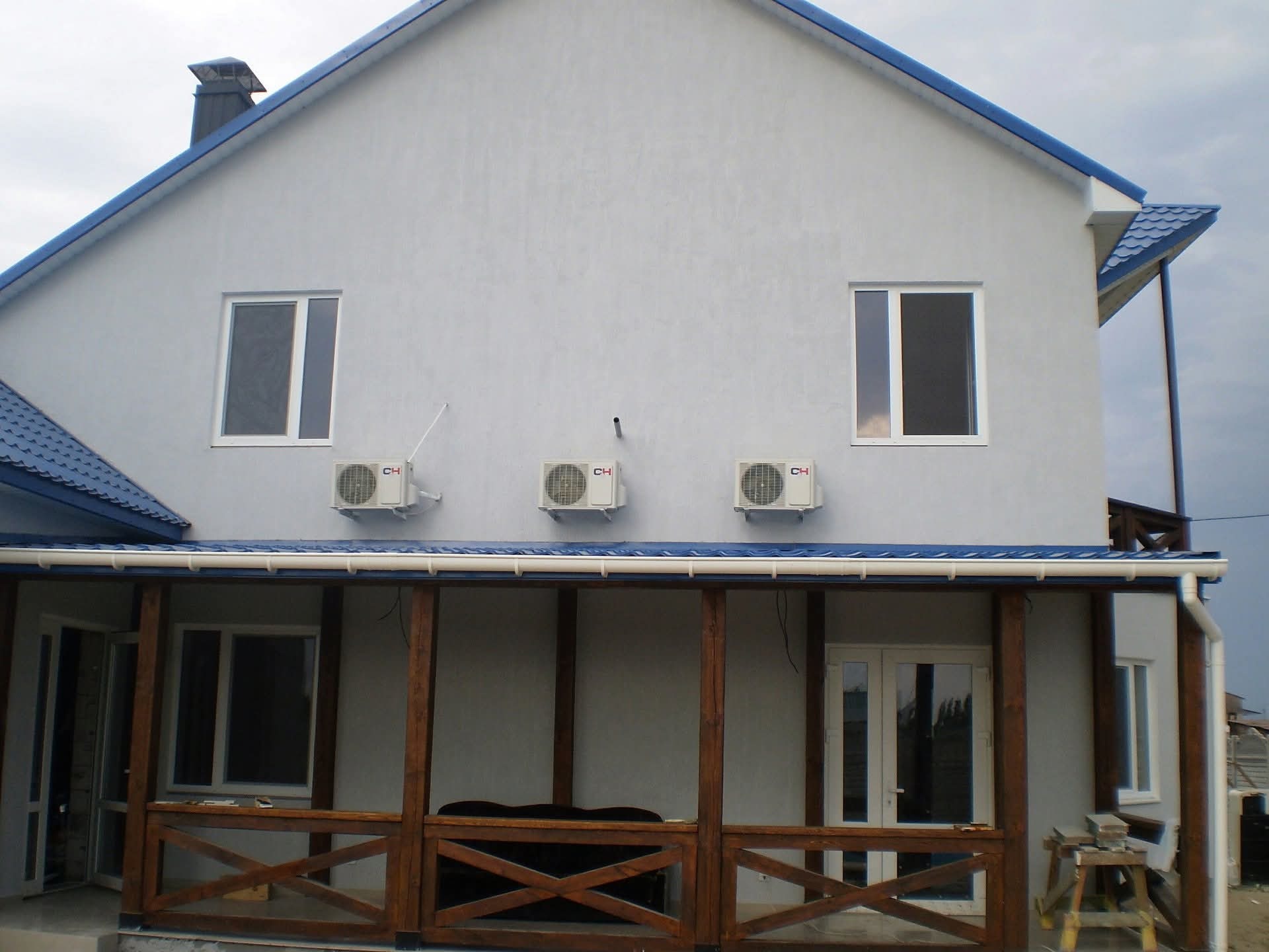 Elewacja domu z szarą fasadą, białymi oknami i trzema klimatyzatorami. Widoczny zadaszony taras z drewnianymi balustradami i niebieski dach.
