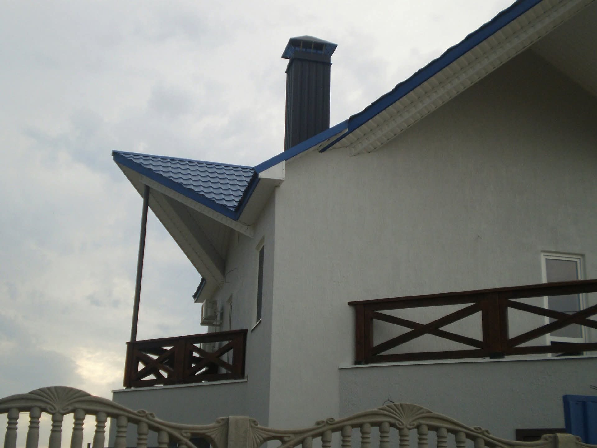Elewacja domu z drewnianą balustradą balkonu i kominem na tle pochmurnego nieba. Widoczna klimatyzacja na ścianie budynku.