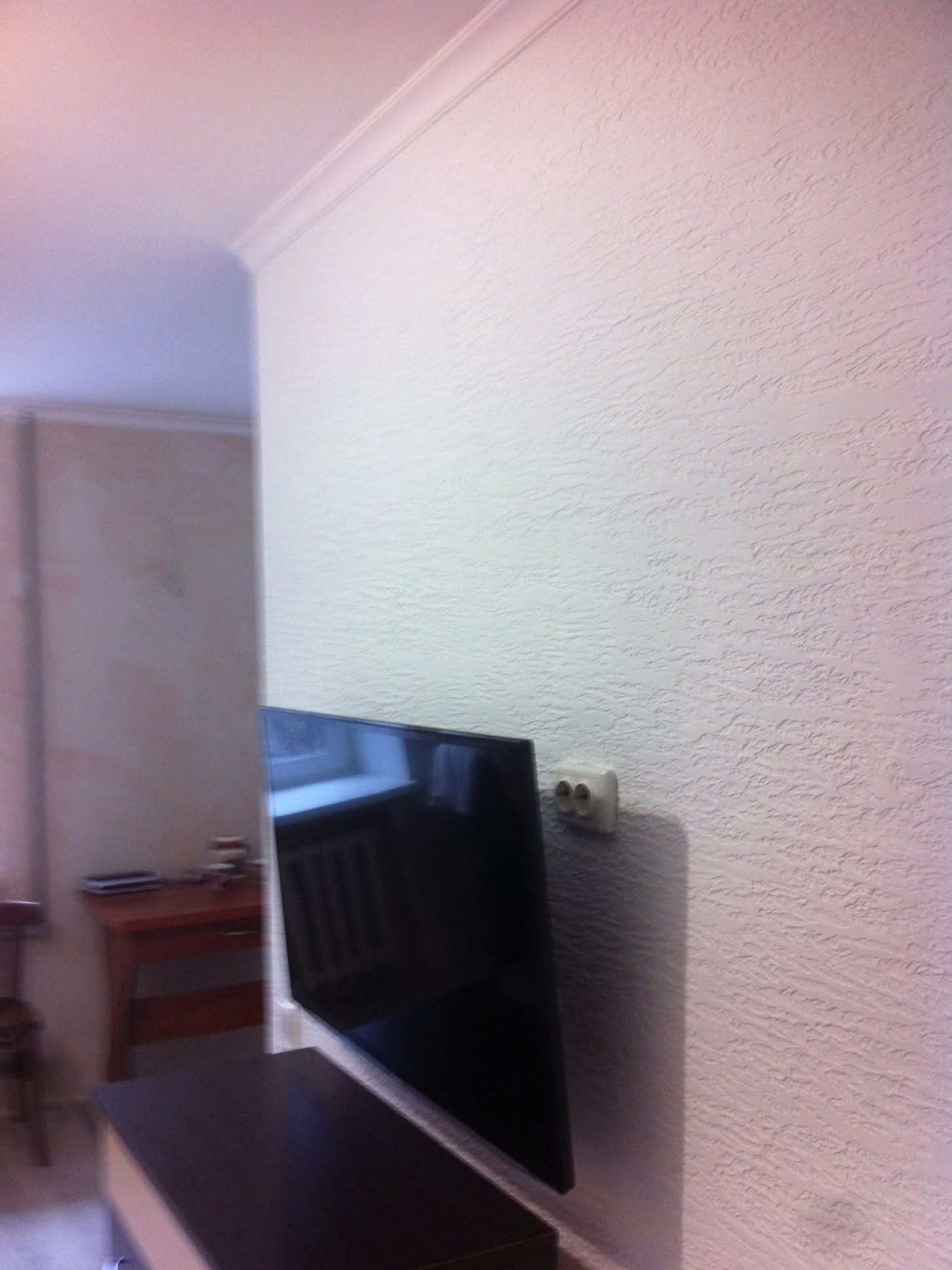Biała ściana z fakturą po szpachlowaniu, widoczny fragment listwy sufitowej i gniazdko elektryczne obok wiszącego telewizora.