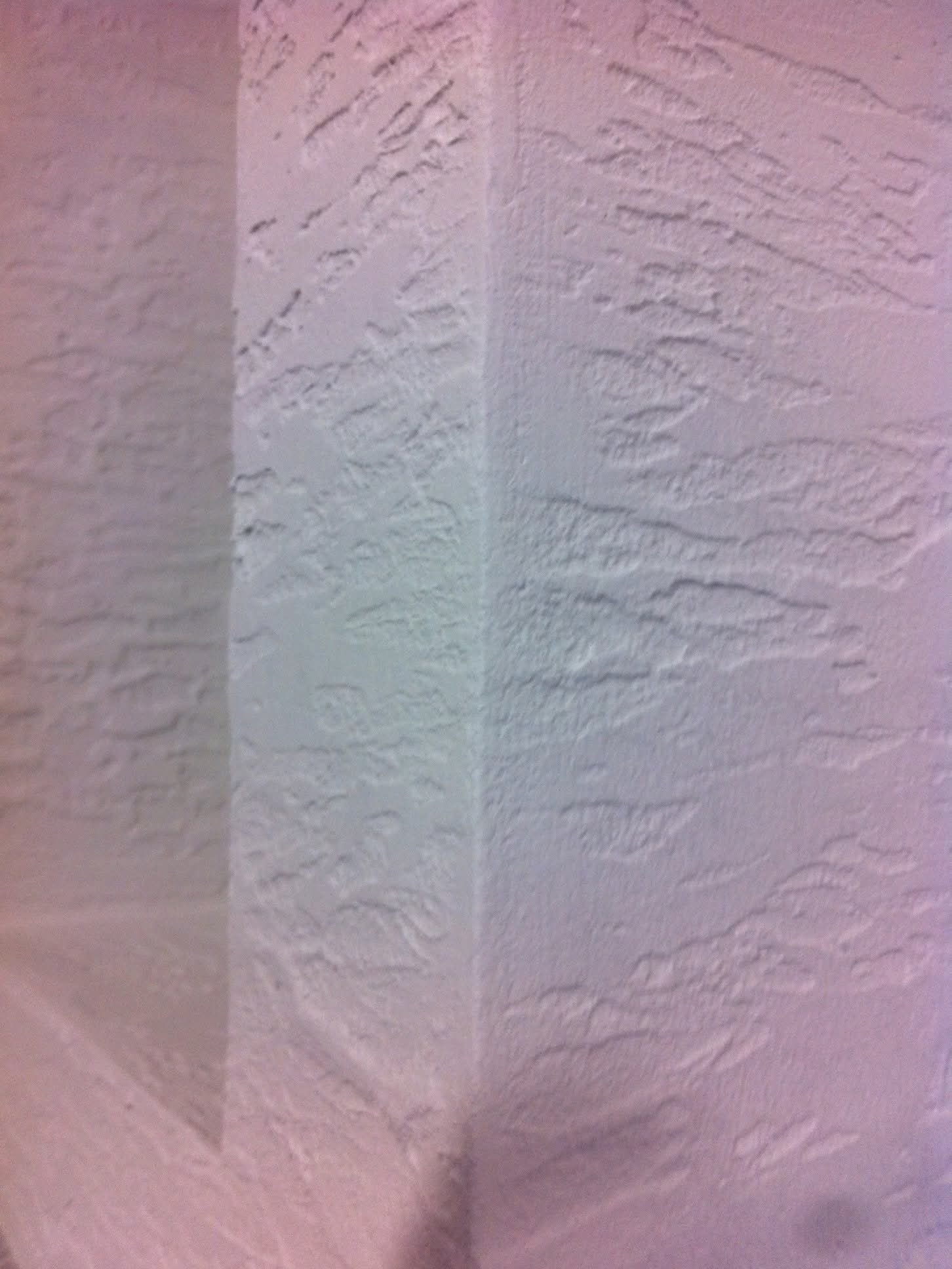 Teksturowana, biała ściana wewnętrzna z widocznym narożnikiem, oświetlona z lewej strony, ukazująca strukturę gładzi w zbliżeniu.