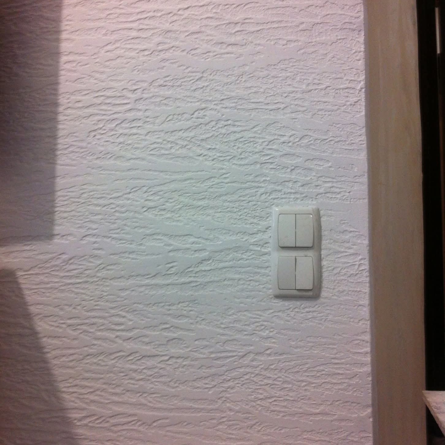 Biała ściana z fakturą, na której zamontowany jest podwójny włącznik światła. Fragment futryny drzwi po prawej stronie. Delikatny cień w lewym górnym rogu.