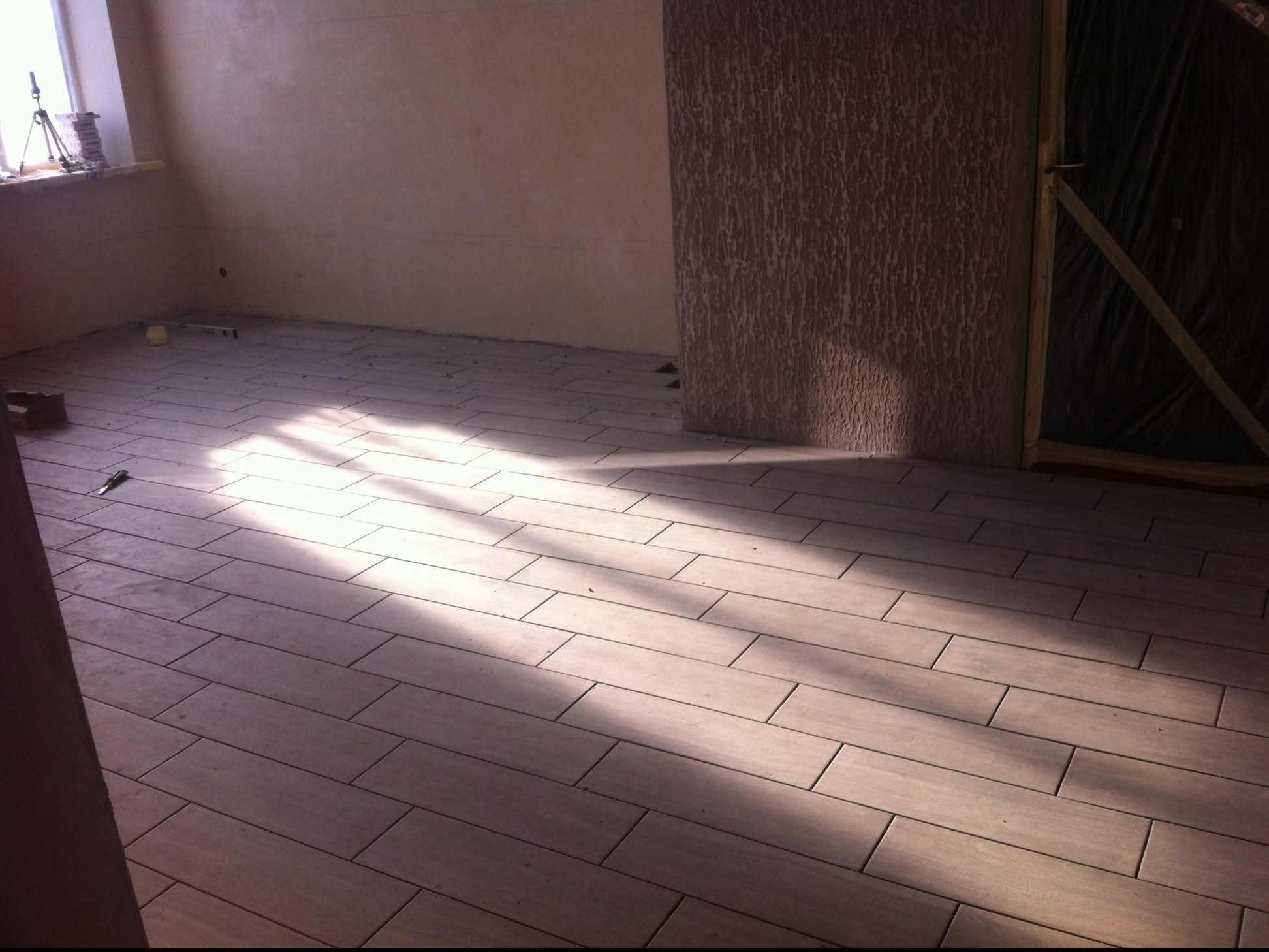 Ułożona podłoga z paneli w kształcie prostokątów, oświetlona promieniami słońca wpadającymi przez okno. Widoczne ściany w odcieniach beżu i fragment okna.