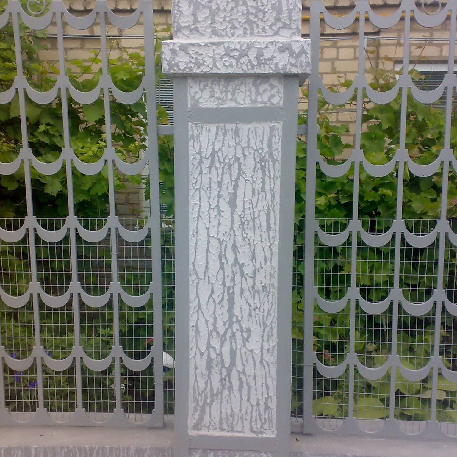 Szary, metalowy płot z ozdobnym wzorem i kolumną o chropowatej fakturze, za którym widać zieleń roślin i fragment muru z cegły.
