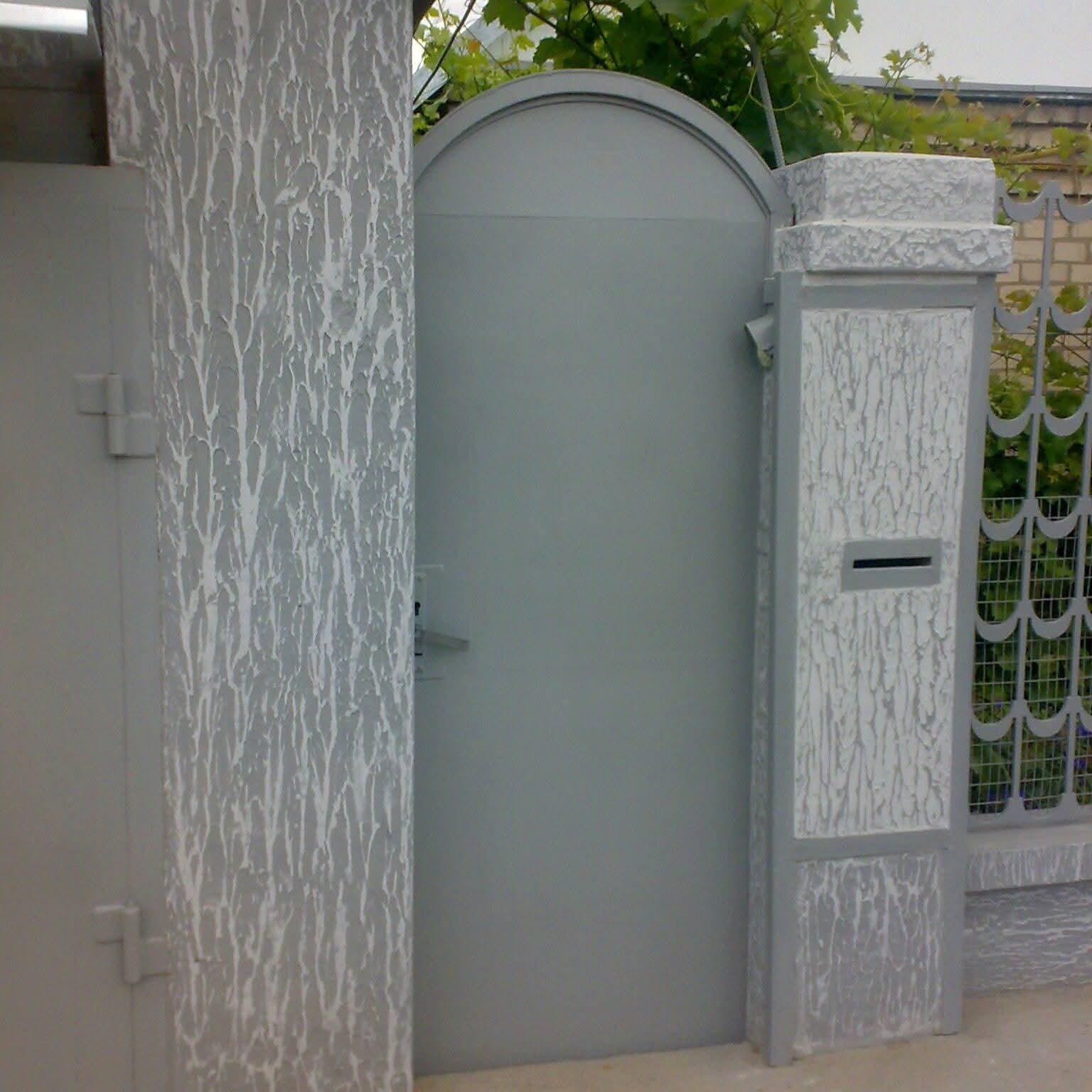 Szara brama z ozdobnymi słupkami i skrzynką na listy, wykończona fakturą imitującą korę drzewa, otoczona zielenią.