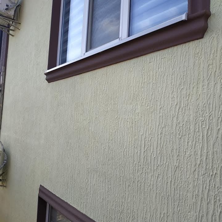 Elewacja budynku z oknem, pokryta jasną, strukturalną farbą o pionowym wzorze. Brązowe obramowanie okien. Widoczny fragment klimatyzatora.