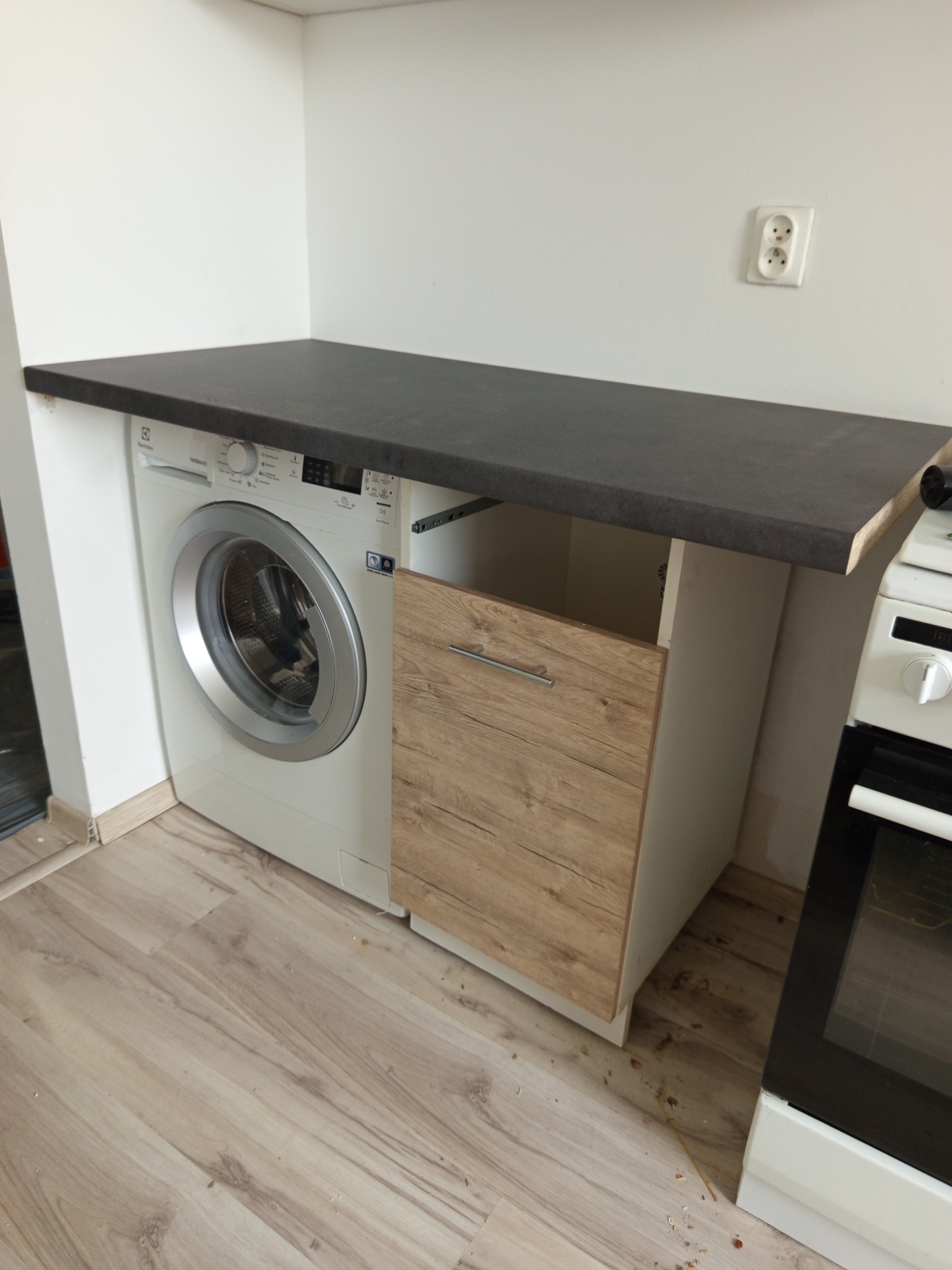Kuchnia: pralka zabudowana pod blatem obok szafki, widok na drewnopodobną podłogę i fragment piekarnika. Gniazdko elektryczne na ścianie.