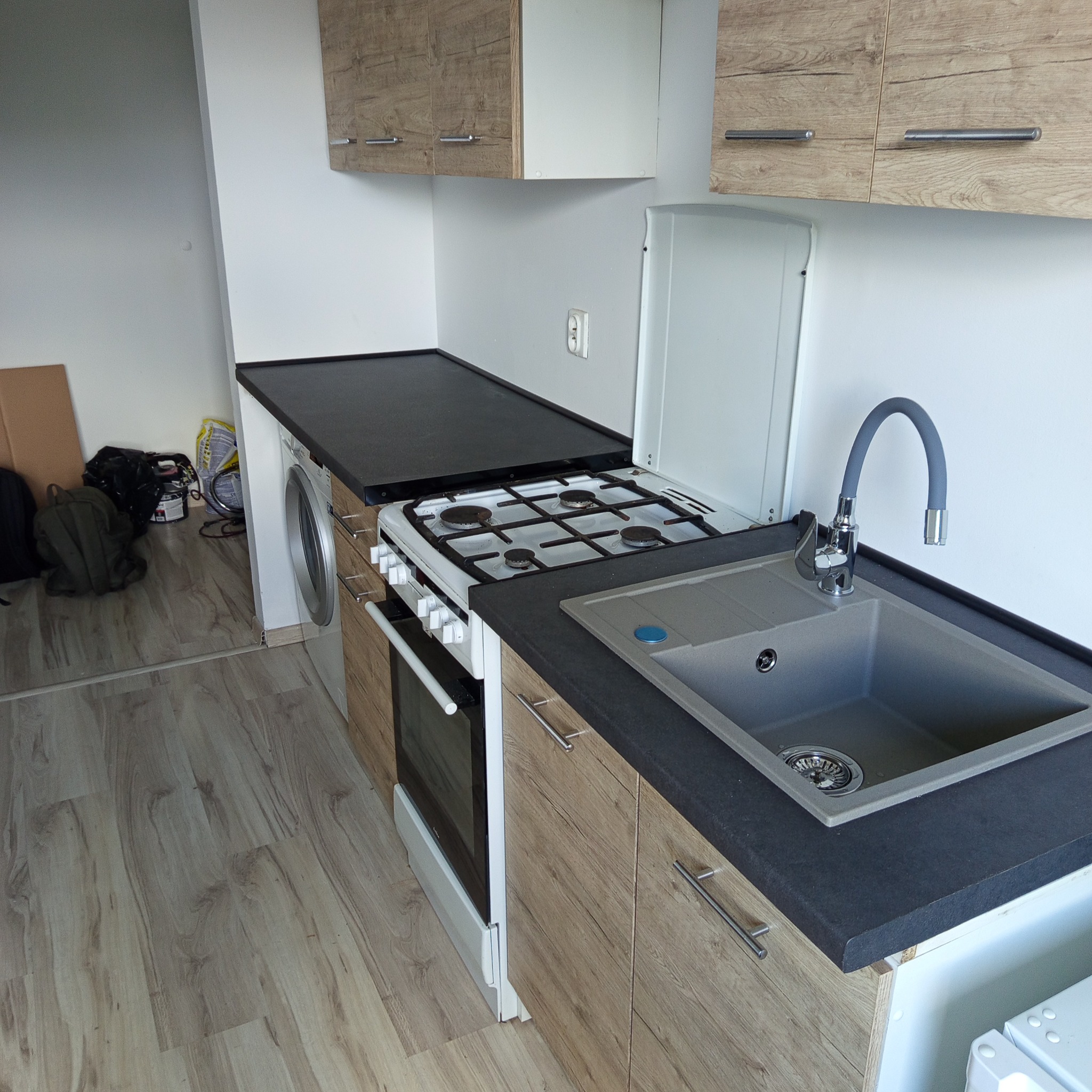 Nowoczesna, kompaktowa kuchnia z drewnianymi szafkami, czarnym blatem i szarą umywalką. Widoczna pralka obok kuchenki. Podłoga z paneli.