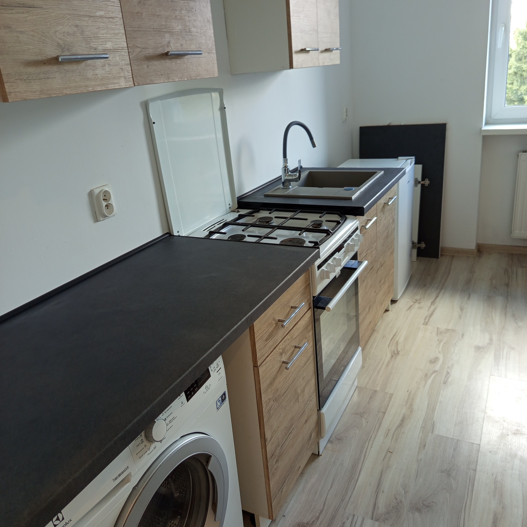 Funkcjonalna, kompaktowa kuchnia z pralką pod blatem, kuchenką gazową i drewnianymi szafkami. Jasne panele podłogowe i okno w tle.