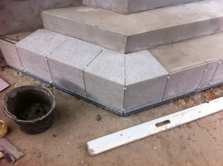 Wykończenie schodów płytkami granitowymi w trakcie prac. Widoczne poziomica, wiadro z zaprawą i krzyżyki dystansowe.