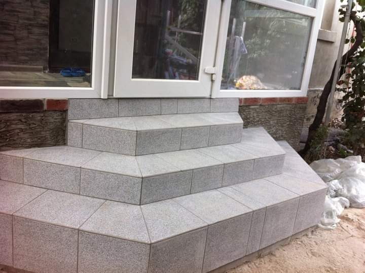 Wykończone płytkami schody wejściowe do domu w Poznaniu. Jasnoszara, drobnoziarnista powierzchnia płytek. Widoczne białe ramy drzwi i okien. Estetyczne detale.