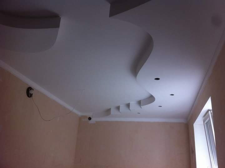 Nowoczesny sufit z płyty gipsowej z falistym wzorem i oświetleniem punktowym. Listwy wykońceniowe przy ścianach. Widok na jasne, beżowe ściany i okno.