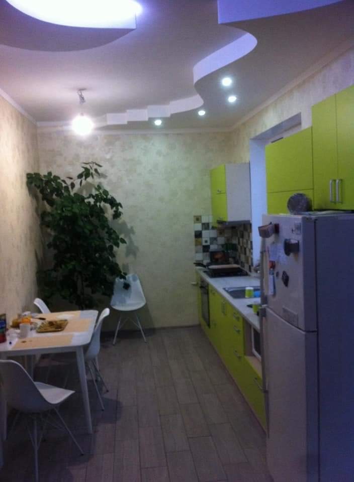 Jasna kuchnia z zielonymi szafkami i dekoracyjnym sufitem. Widoczne panele podłogowe, stół z krzesłami i roślina doniczkowa w rogu pomieszczenia.