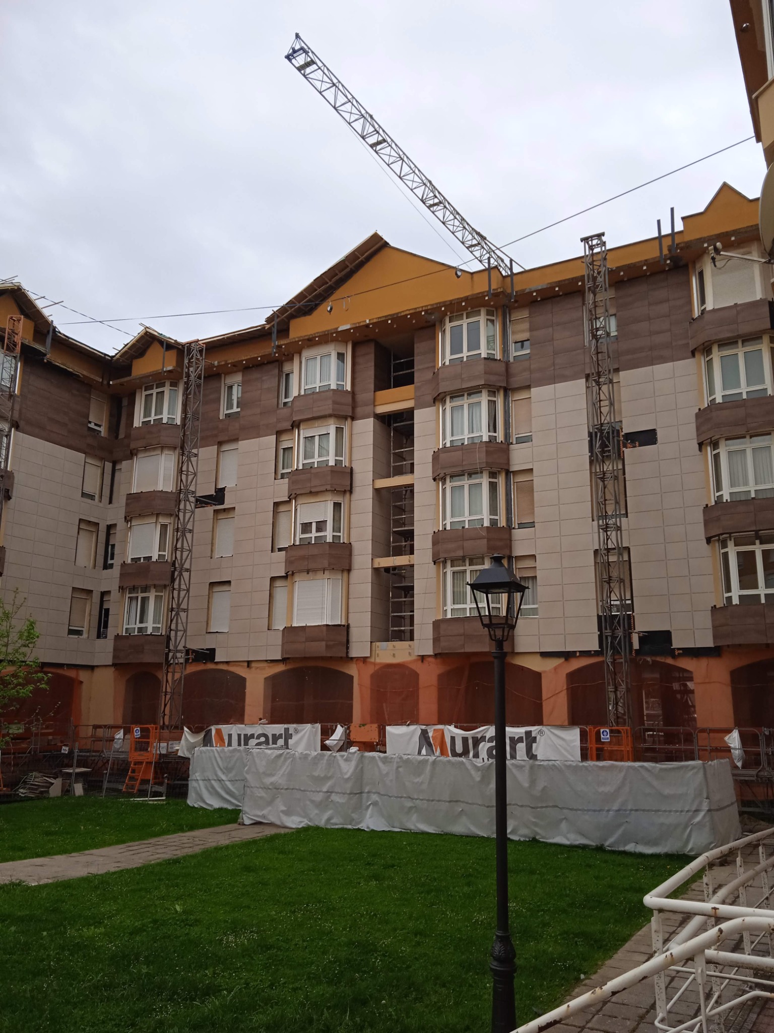 Wielopiętrowy budynek w trakcie remontu elewacji z widocznym dźwigiem i rusztowaniami. Prace zabezpieczone płachtą z logo. Widok z zewnątrz na teren zielony.