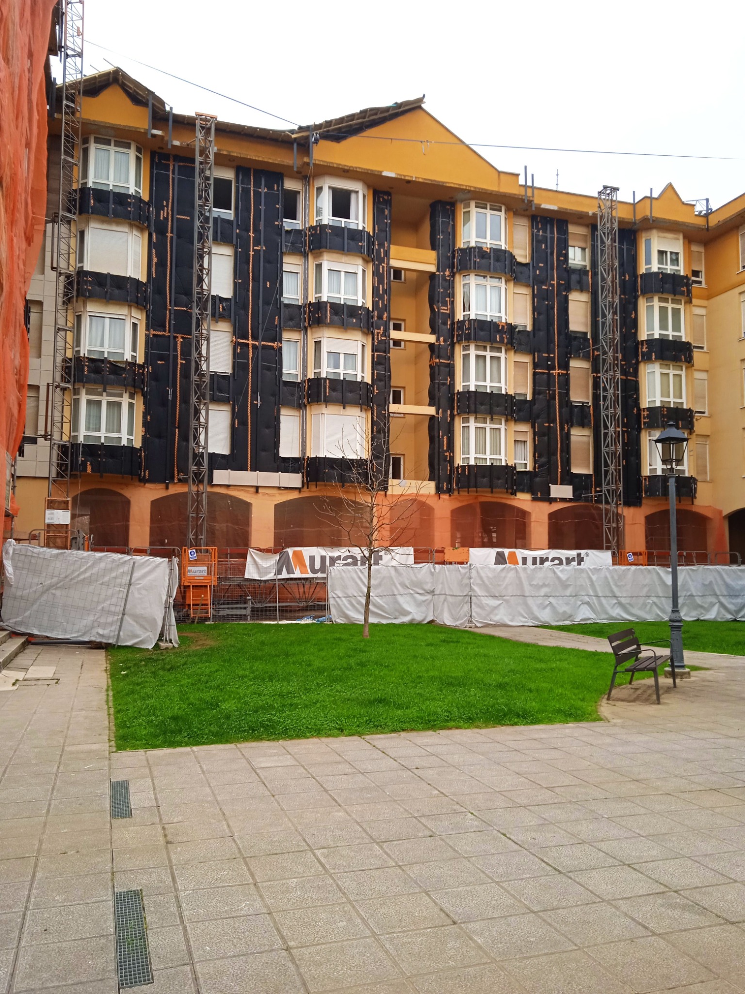 Ocieplanie budynku wielorodzinnego z zamontowanym rusztowaniem i siatką zabezpieczającą. Widoczne płyty izolacyjne na elewacji w kolorze czarnym.