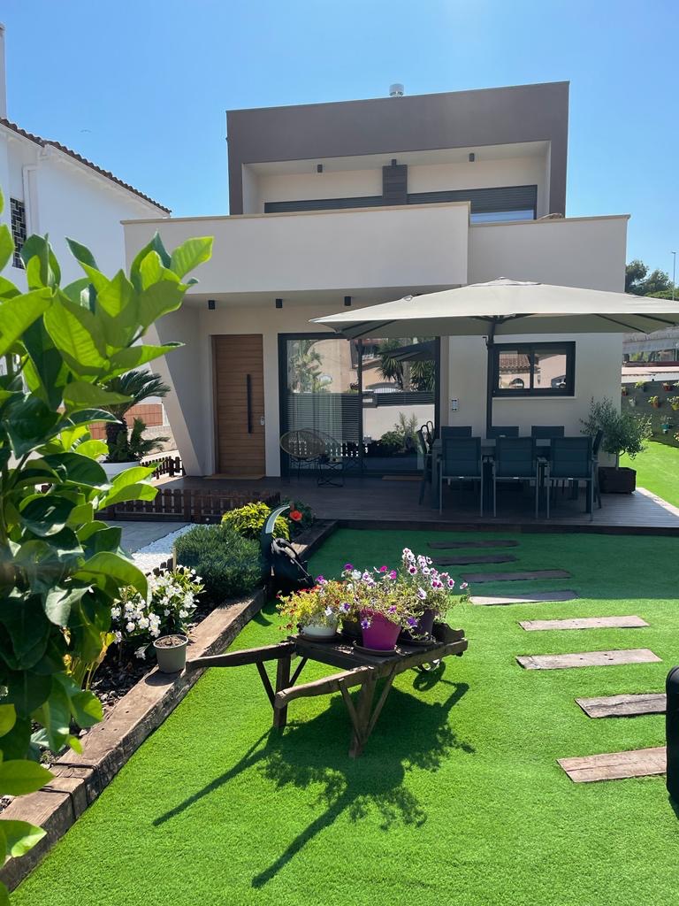 Nowoczesny dom z ogrodem: drewniany taras, parasol, wózek z kwiatami na trawniku. Minimalistyczna bryła budynku z płaskim dachem i dużymi przeszkleniami.