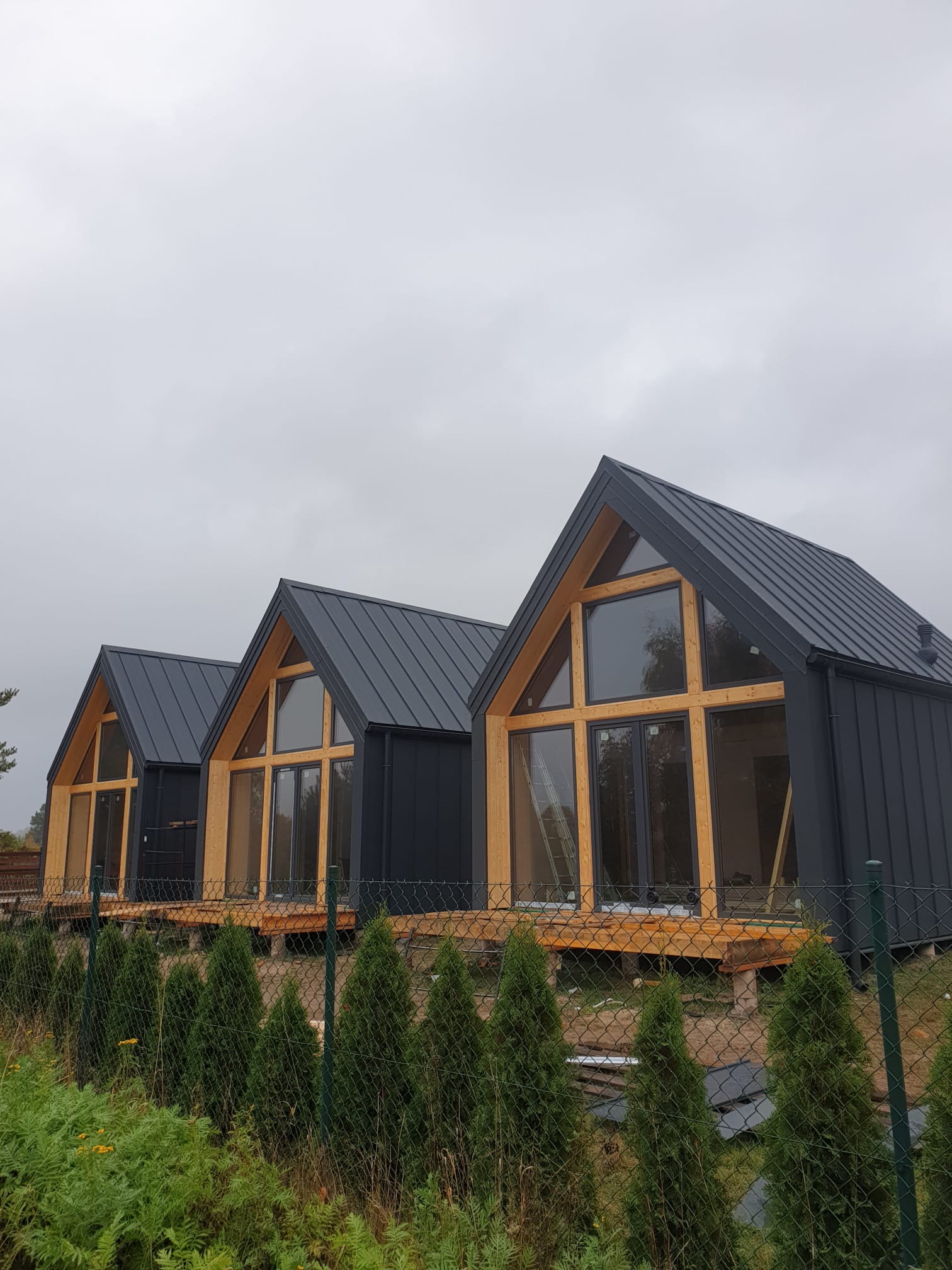 Trzy identyczne, nowoczesne domki szkieletowe z ciemną blachą na dachu i dużymi oknami, ustawione w rzędzie na tle pochmurnego nieba, widoczne zza ogrodzenia.