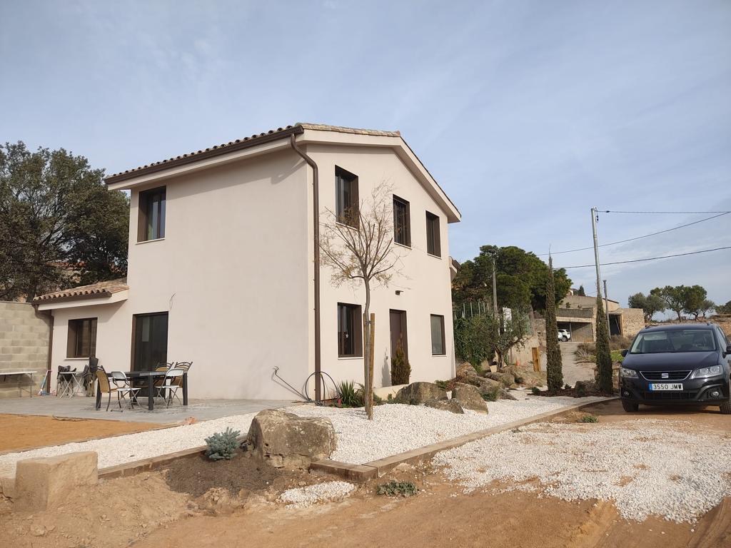 Dwukondygnacyjny dom z beżową elewacją i ciemnymi oknami. Biały żwir wokół, auto zaparkowane obok. Widok z zewnątrz w słoneczny dzień.