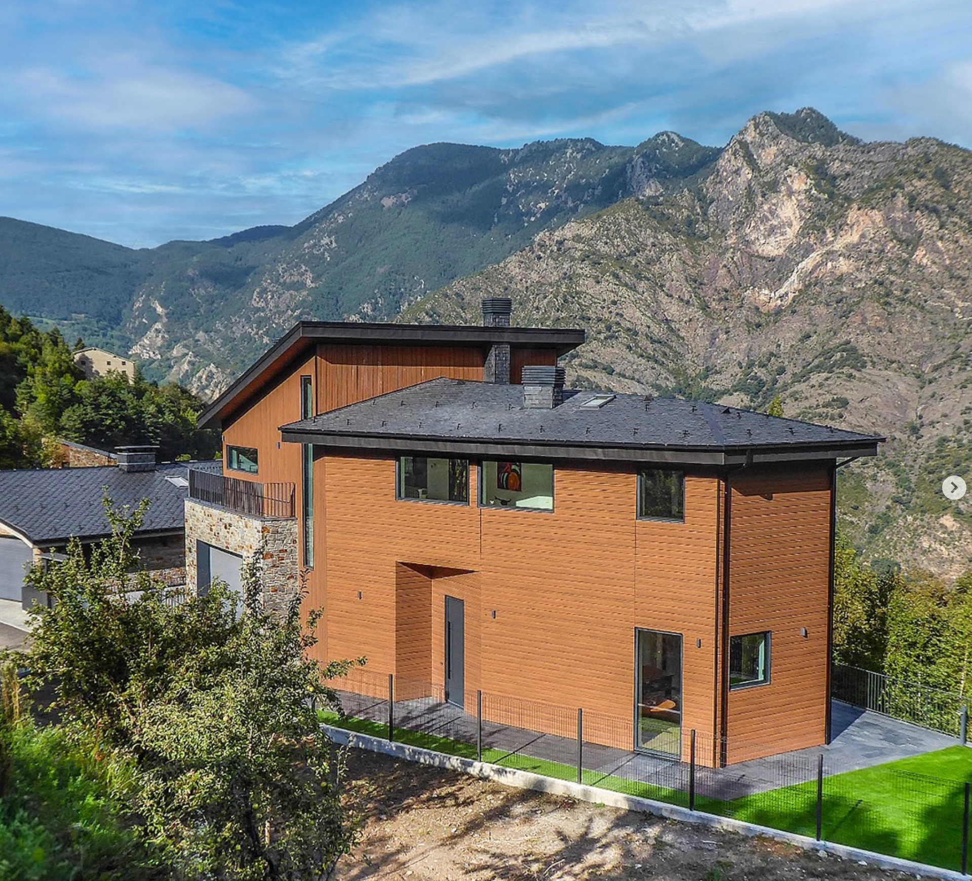 Dwukondygnacyjny dom szkieletowy z drewnianą elewacją na tle gór, z kamiennym wykończeniem balkonu i czarnym dachem. Widok z lekkiej góry.