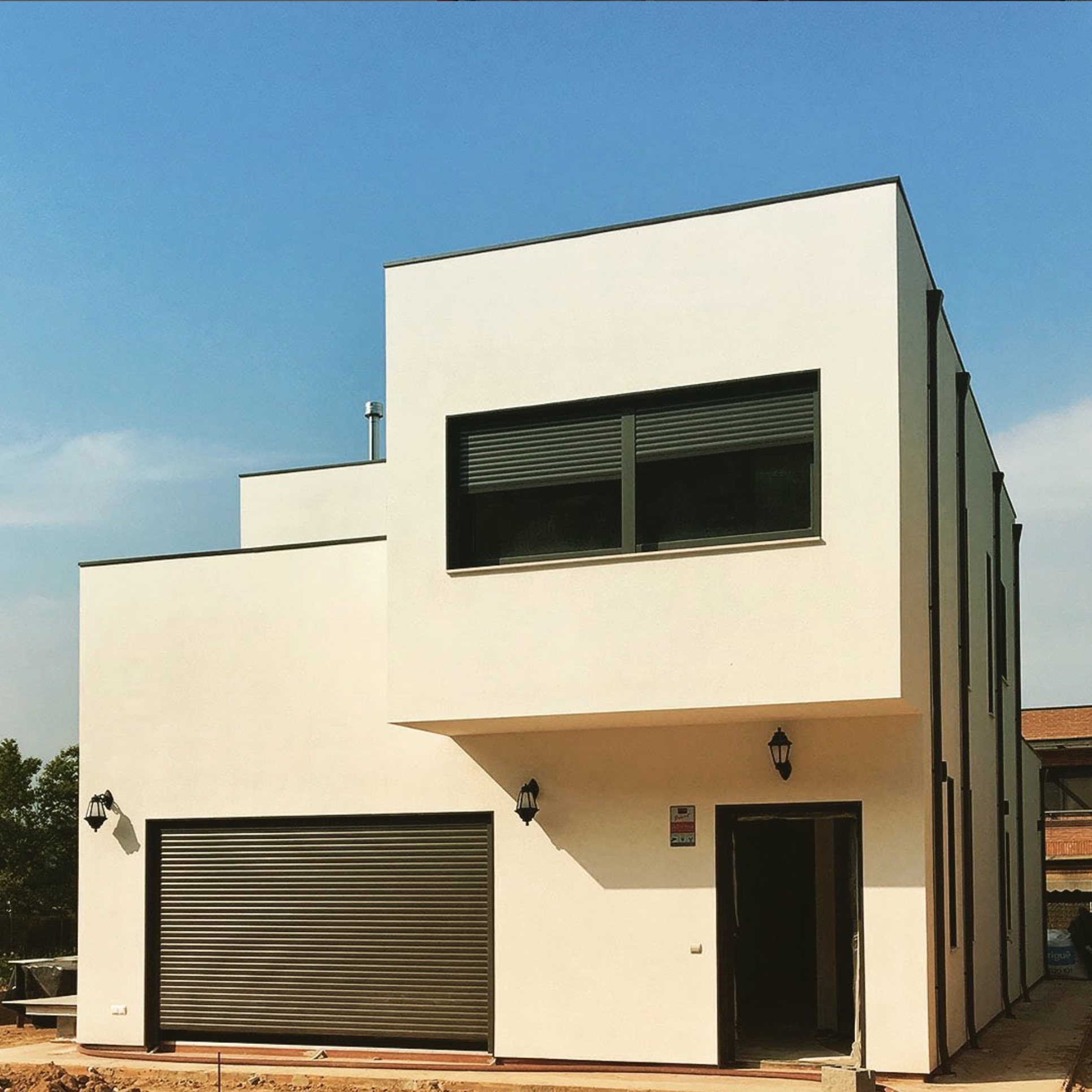 Nowoczesny, dwukondygnacyjny dom o prostej bryle, z garażem zamykanym roletą i oknem z żaluzjami, w jasnej kolorystyce, na tle błękitnego nieba.