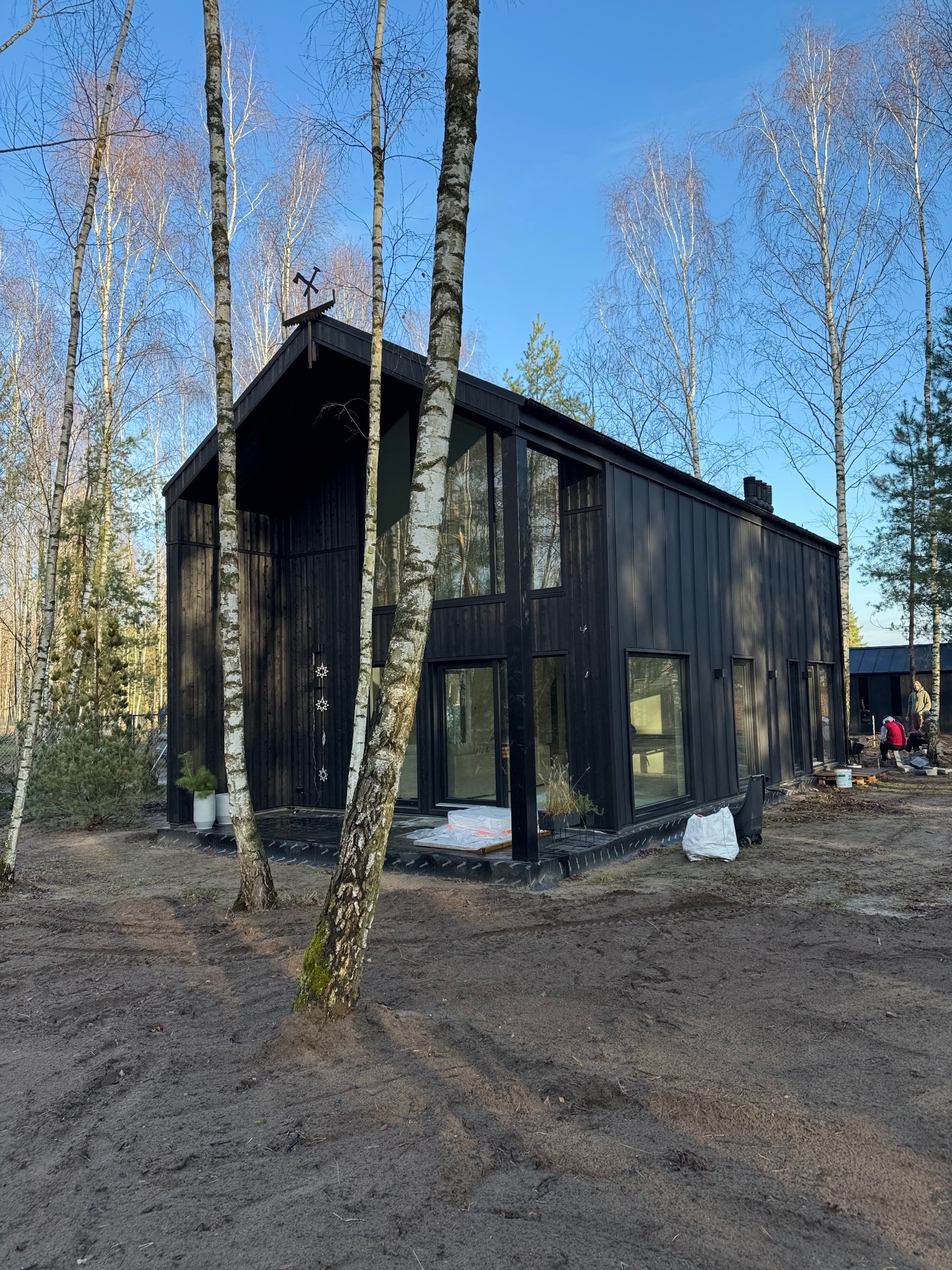 Czarny dom szkieletowy z dużymi oknami, otoczony brzozami i przygotowanym terenem. Na dachu dekoracyjny element w kształcie skrzyżowanych narzędzi.