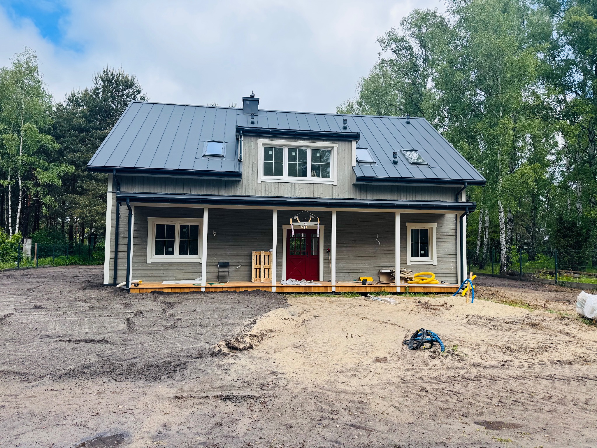 Szary dom z drewnianą elewacją i ciemnoszarym dachem, czerwone drzwi wejściowe, widok z zewnątrz na tle drzew, teren przygotowany pod zagospodarowanie.