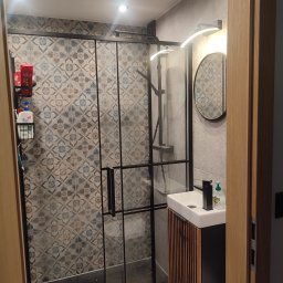 Emsi Expert & Home - Nowoczesna łazienka z kabiną prysznicową typu walk-in, wzorzyste płytki, umywalka z szafką z lamelami i okrągłe lustro z oświetleniem LED.