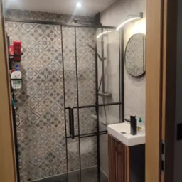 Nowoczesna łazienka z kabiną prysznicową typu walk-in, wzorzyste płytki, umywalka z szafką z lamelami i okrągłe lustro z oświetleniem LED.