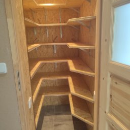 Emsi Expert & Home - Wykonana z płyty OSB spiżarnia z półkami, oświetlona punktowym światłem. Widoczne metalowe wsporniki półek i wykończenie z surowego drewna. Podłoga wyłożona płytkami.